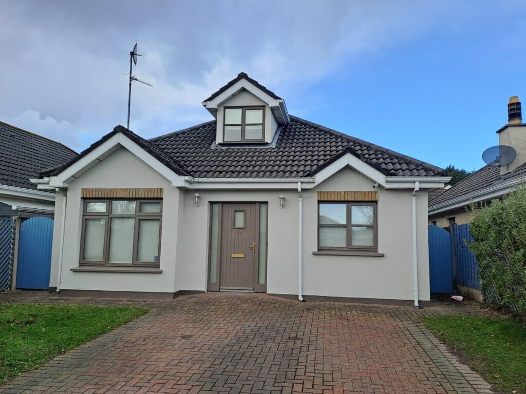 A modern detached bungalow with a grey tiled roof and a dormer window, featuring a freshly painted light grey exterior with white trim. The house has two symmetrical front windows with brown accents and a central wooden front door with a brass letterbox and handle. A paved driveway leads up to the entrance, flanked by a small lawn and a blue wooden fence on both sides. The background features a partly cloudy sky and neighbouring houses with satellite dishes and chimneys
